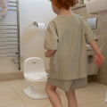 A young child with curly red hair stands in a bathroom facing the At Home and On the Go Potty Bundle with Book, placed near a toilet, with a towel rack and toilet paper holder mounted on the wall.