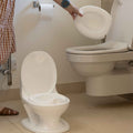 A person empties the My Real Mini Toilet Potty into a regular toilet, standing by a wall-mounted toilet paper roll—wearing a brown plaid dress and showing an arm tattoo—while helping with potty training cleanup.
