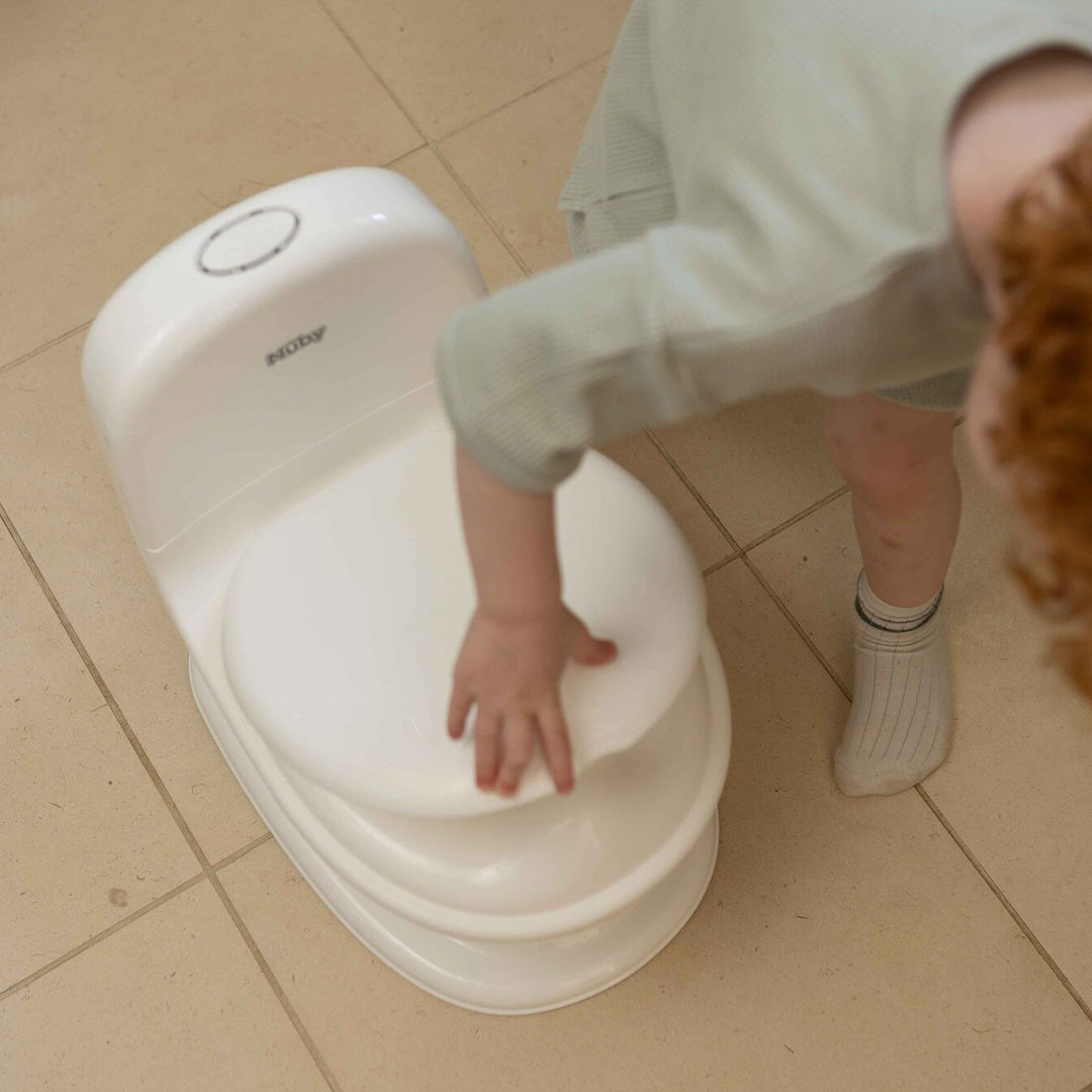 A young child with curly hair touches the lid of a My Real Mini Toilet Potty on a tiled floor, only showing one arm and part of their body.