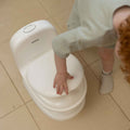 A young child with curly hair touches the lid of a My Real Mini Toilet Potty on a tiled floor, only showing one arm and part of their body.