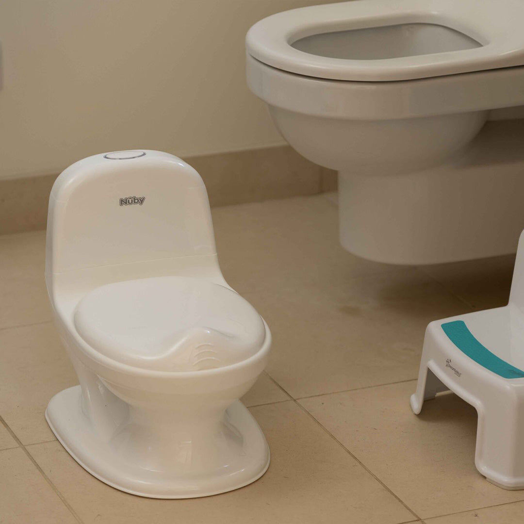The My Real Mini Toilet Potty, a white plastic potty for training, is placed on a tiled bathroom floor next to a wall-mounted toilet and a small step stool with a teal top.