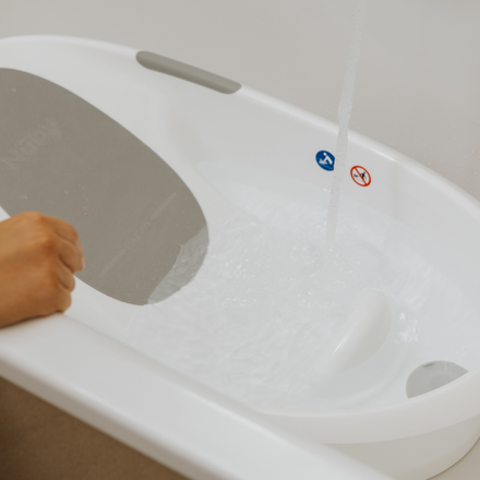 A hand holds a gray panel over a white bathtub as water fills it, with the Nuby UK Newborn Bath & Thermometer Bundle and two safety stickers on the rim for a safe baby bath.