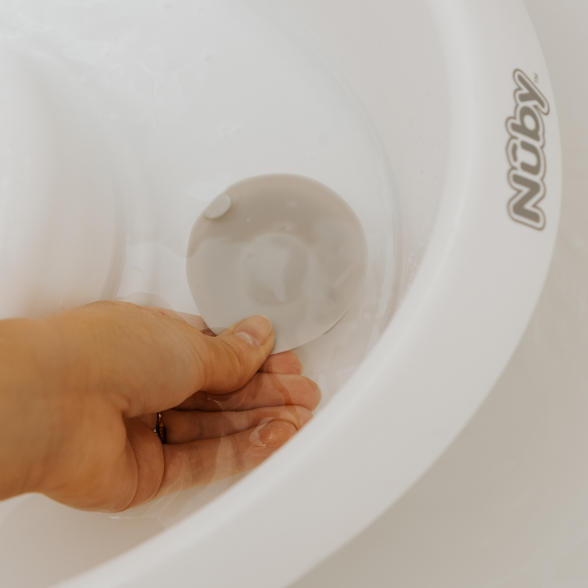 A hand touches a silicone suction cup at the bottom of a white Nuby UK Newborn Bath & Thermometer Bundle, perfect for baby bath time. The basin features the Nuby logo on the rim.