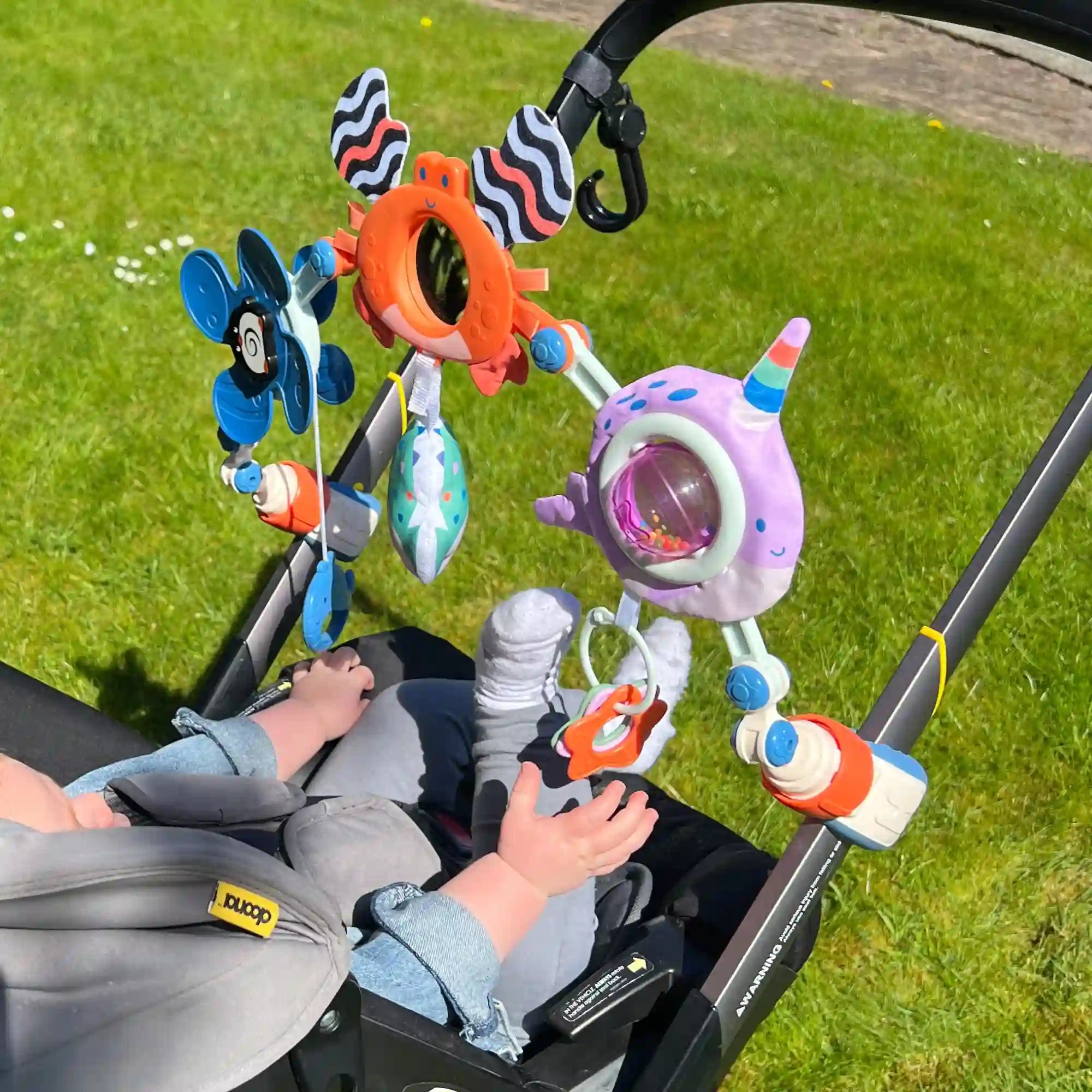 Ocean Friends Pram Toy Arch. A baby reaching to play with their Nuby pram arch toy in an outdoor setting.