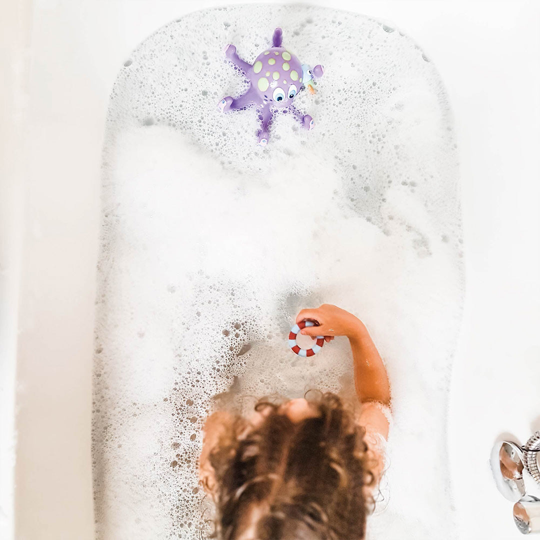 A young child with curly hair plays in a bubble bath, holding a toy life preserver while a purple octopus from the Nuby UK Bath Toy Bundle 12 Months+ floats nearby.