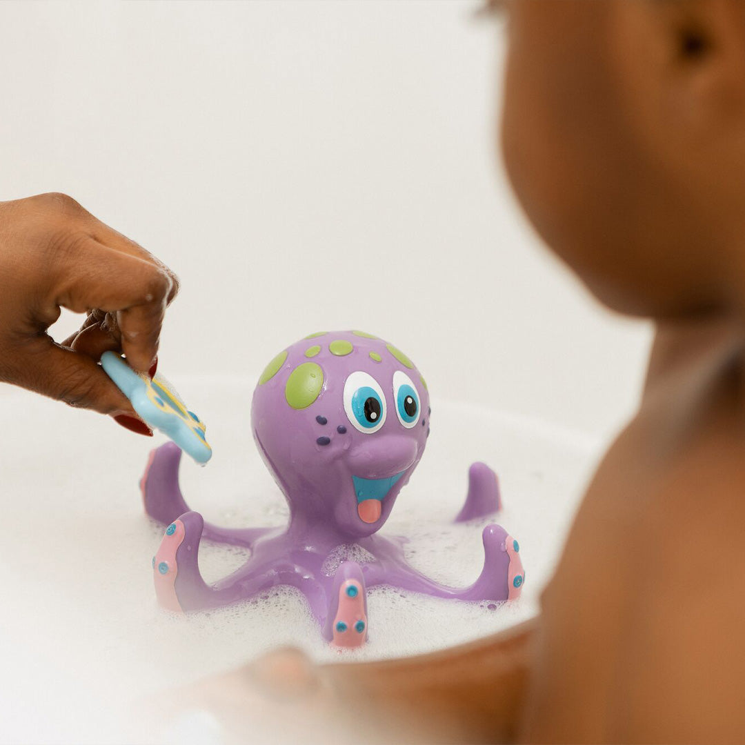 A child enjoys bubbly bath time, gazing at a Nuby UK Bath Toy Bundle 12 Months+ octopus while an adult holds another blue toy, making bathtime playful and sensory with these charming Nuby UK bath toys.