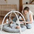 A woman smiles at a baby lying under the Nuby Penguin and Pals Play Gym with hanging toys in a bright, cozy nursery, with a crib and dresser in the background.