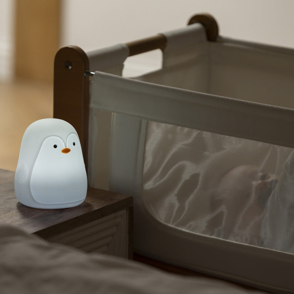 The Penguin Colour Changing Night Light glows softly on a bedside table beside a crib with sheer mesh sides, where a baby is partially visible inside.