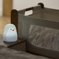 The Penguin Colour Changing Night Light glows softly on a bedside table beside a crib with sheer mesh sides, where a baby is partially visible inside.