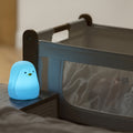 A Penguin Colour Changing Night Light sits on a table beside a bassinet with a sleeping baby partially visible through the mesh, filling the peaceful scene with its gentle, soothing glow.