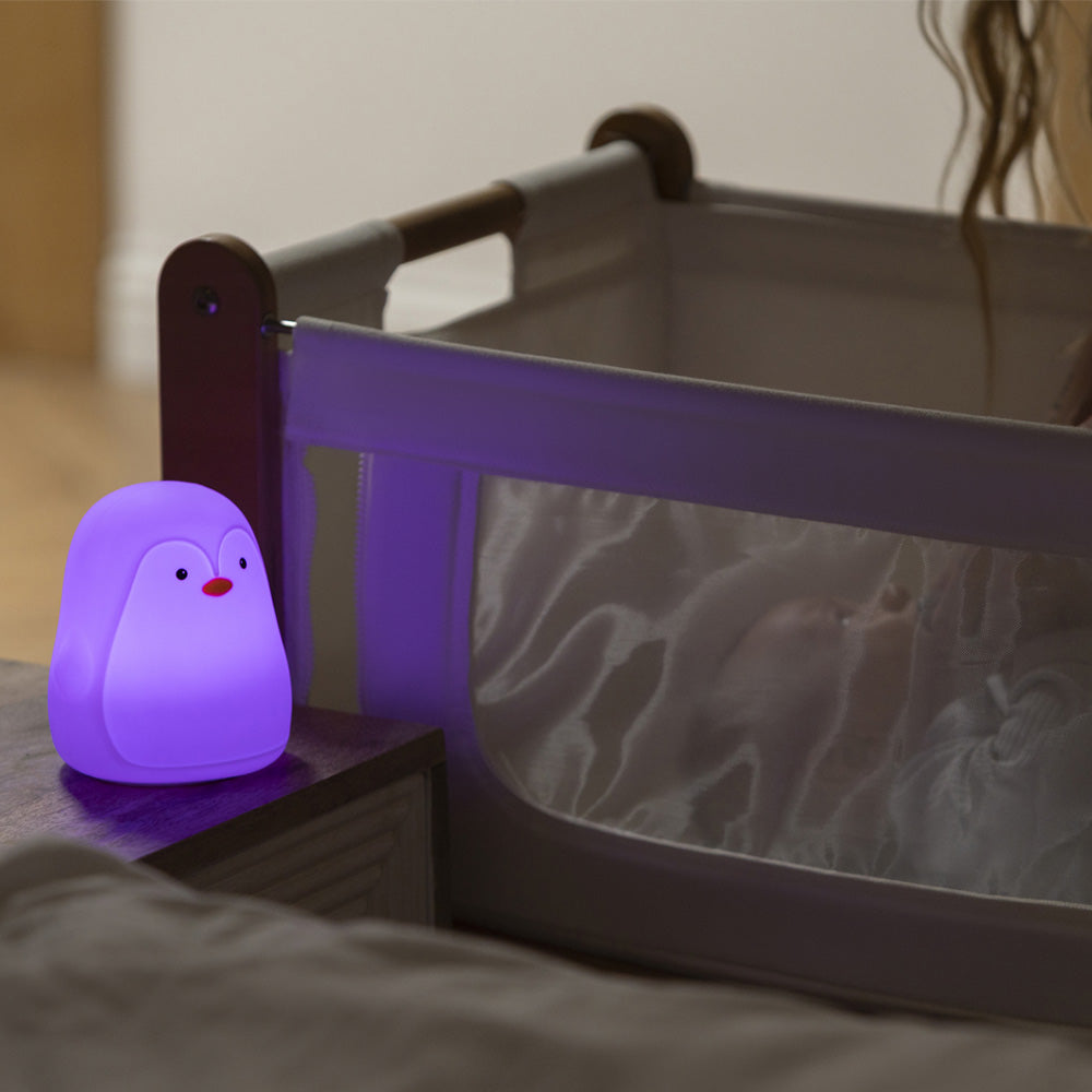 The Penguin Colour Changing Night Light glows gently on a bedside table by a crib with mesh sides, casting a soothing light over the sleeping baby resting under a light blanket.