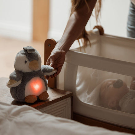 A person places the Nuby Penguin Sleep Aid with Cry Sensor, which has a glowing orange baby soothing light, near a sleeping baby in a crib with a pacifier.