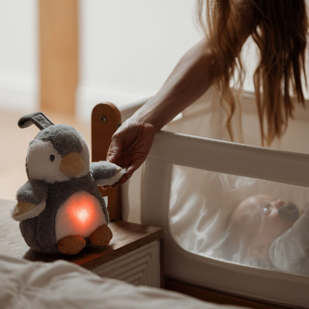 A person places the Nuby Penguin Sleep Aid with Cry Sensor, featuring a red soothing light, near a baby lying in a crib partially covered by a mesh side panel.