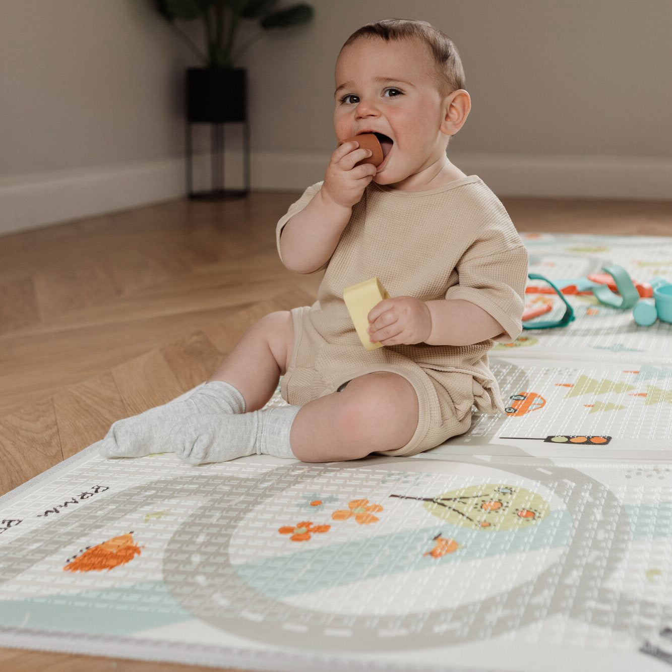 A baby sat up while playing on the Play Mat decorated in a road pattern including cars trees and flowers.