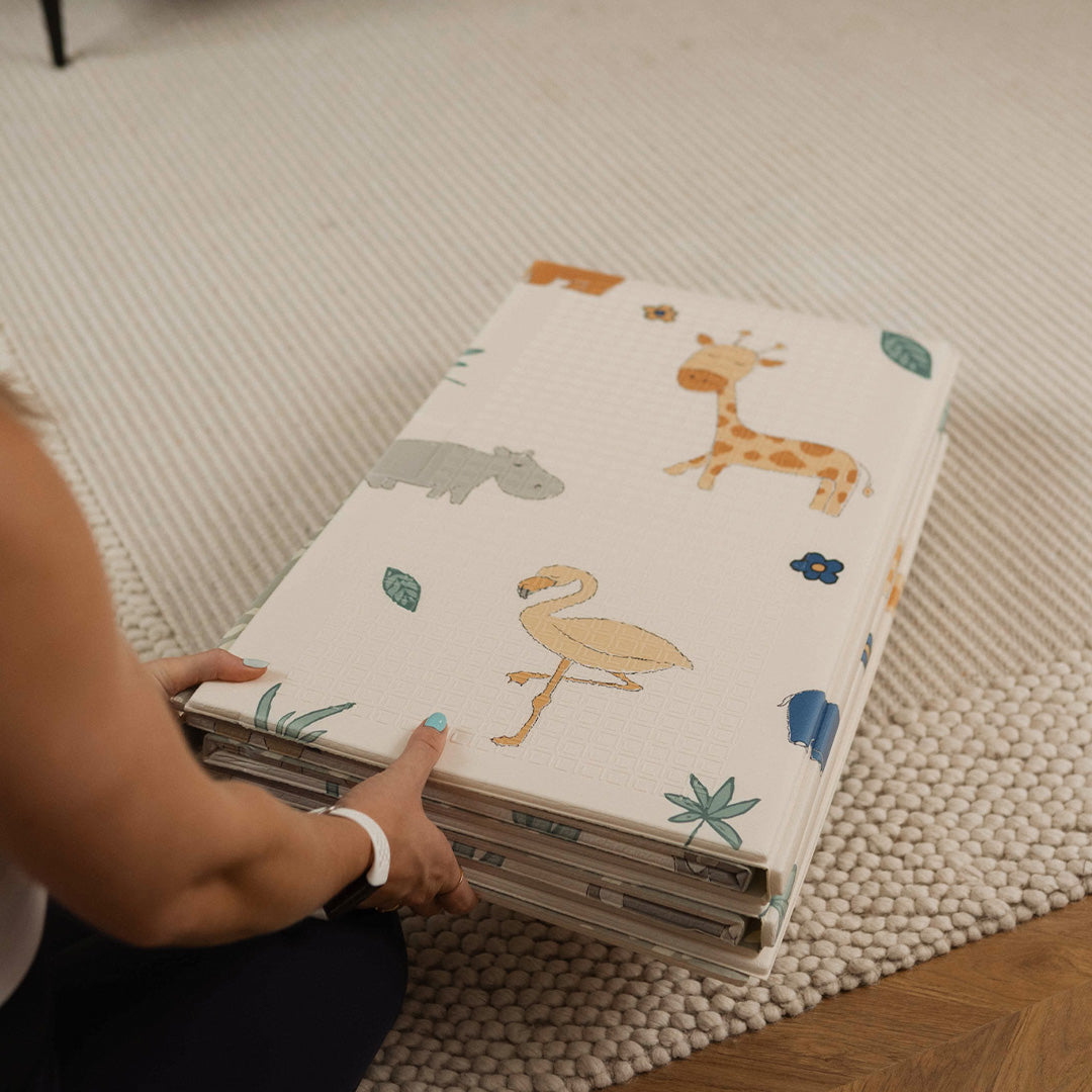 A person wearing a white wristband holds the Nuby Foldable Play Mat, Safari, featuring colorful animal illustrations like a giraffe, hippo, and flamingo—ideal as a reversible foam mat or for tummy time on textured cream carpet.