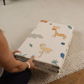 A person wearing a white wristband holds the Nuby Foldable Play Mat, Safari, featuring colorful animal illustrations like a giraffe, hippo, and flamingo—ideal as a reversible foam mat or for tummy time on textured cream carpet.