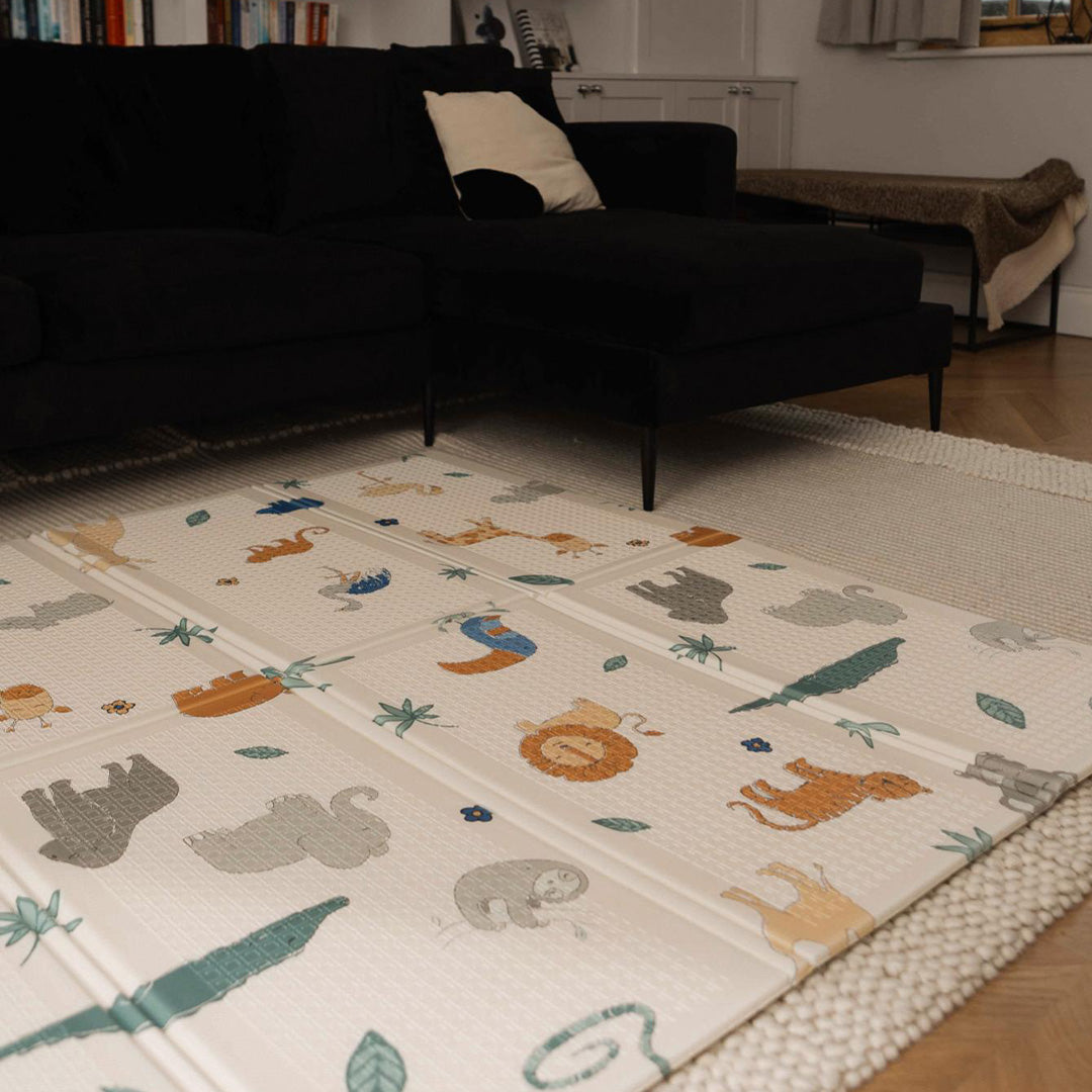 The Nuby Foldable Play Mat, Safari, featuring colorful animal designs, is spread out on the floor of a cozy living room with a dark sofa and neutral rug.