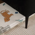 The Nuby Foldable Play Mat, Safari, featuring a cartoon tiger and leaves, is partially tucked under a black sofa with slender legs beside a textured beige carpet.