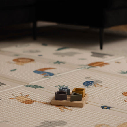 A Nuby Foldable Play Mat, Safari, featuring animal illustrations, lies beneath a wooden shape sorter toy. The patterned mat is reversible, and part of a dark-colored couch can be seen in the background.