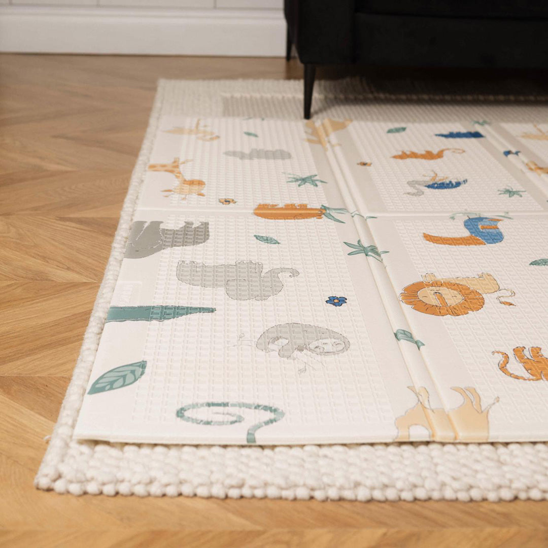 The Nuby Foldable Play Mat, Safari, featuring colorful animal illustrations like a lion and sloth, rests on a textured cream rug atop a wooden floor near a dark sofa.