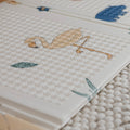 A close-up of the Nuby Foldable Play Mat, Safari, shows its reversible foam design with a textured white surface featuring a simple flamingo on one leg and green leaf patterns, placed on a woven beige surface for style and comfort.