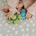 A baby in a pink sweater sits on a patterned blanket, holding the Nuby Pop-it Teether Toy 2 Pack—one shaped like a turtle and the other like a blue fish—designed as sensory toys for babies.