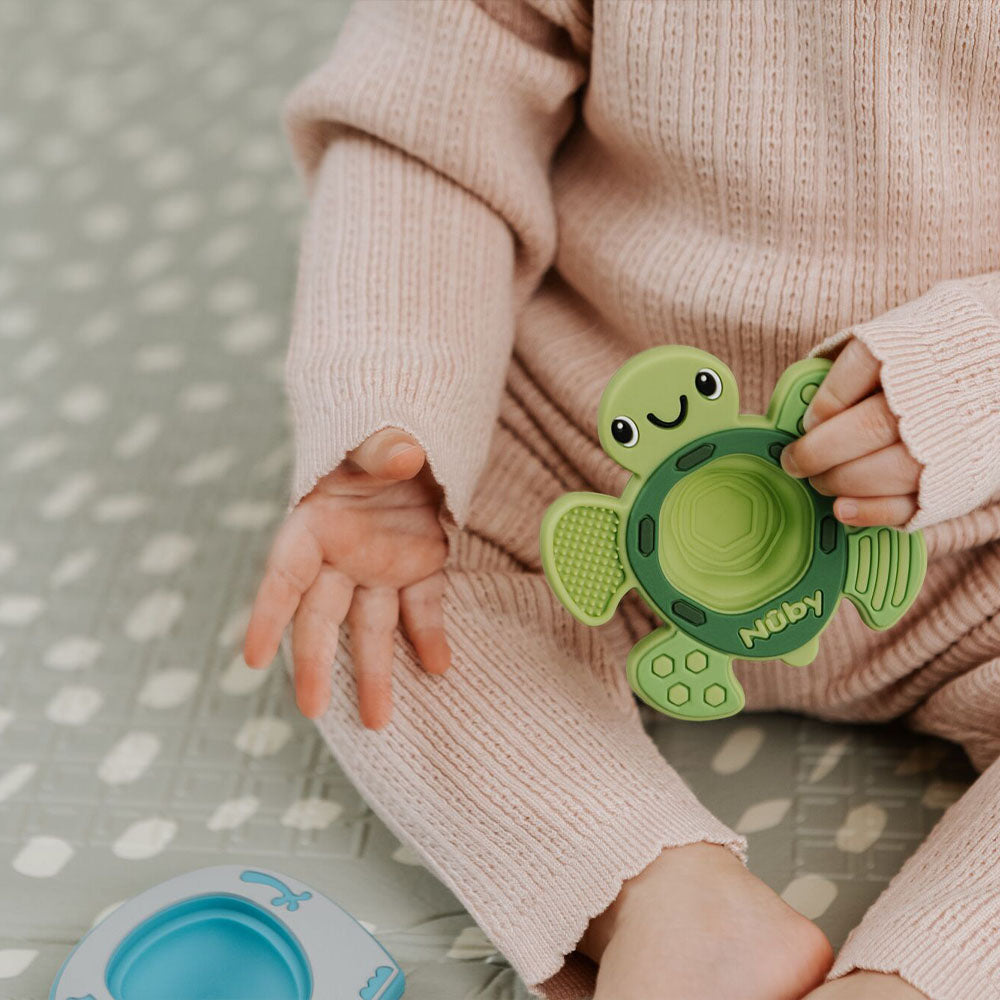 A baby in a pink knit outfit sits on a patterned blanket, holding the green turtle-shaped Pop-it Teether Toy from Nuby. Nearby, part of a blue Nuby sensory toy for babies is also visible.