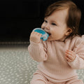 A brown-haired baby in a pink knitted outfit sits on a patterned mat, holding and chewing on a Nuby Pop-it Teether Toy 2 Pack in blue, shaped like a whale.