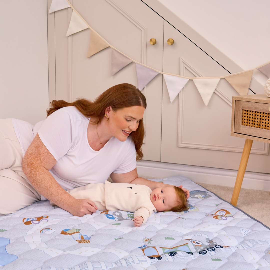 A woman with long red hair smiles at a baby lying on the Quilted Playmat indoors, beside a small wooden table and a wall decorated with beige and white bunting.