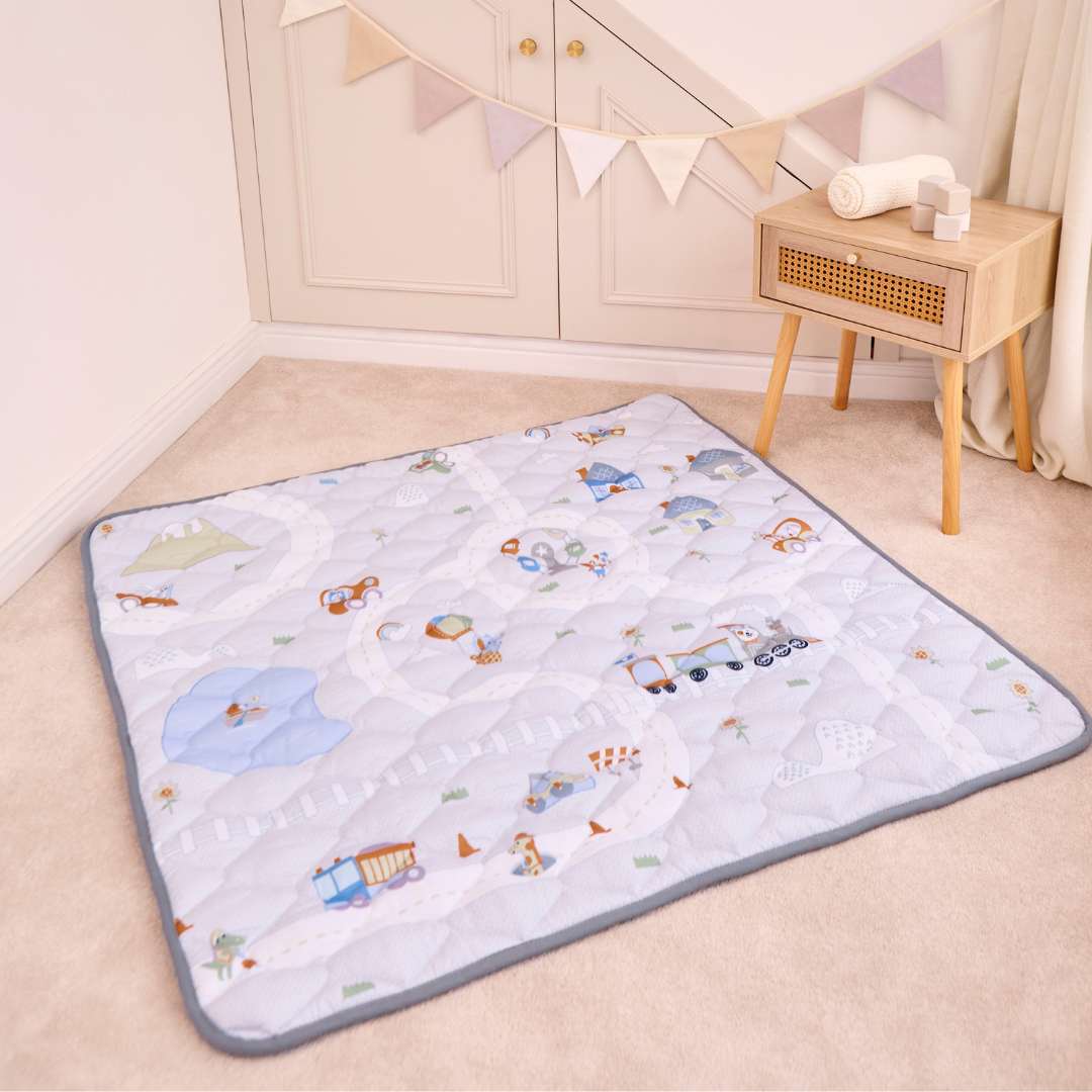 The Quilted Playmat, featuring cheerful vehicle and animal designs, rests on a beige carpet. Next to it stands a wooden side table with towels, while colorful decorative bunting hangs above.