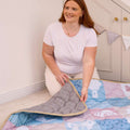 A smiling woman with long red hair and a white shirt kneels in a bright room with bunting and cabinets, spreading out the Quilted Playmat.