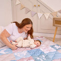 A smiling woman with long red hair and a white shirt lies on a Quilted Playmat, holding her baby who rests on their back. Pastel bunting decorates a cabinet in the background.