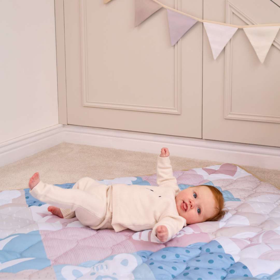 A baby in a light outfit smiles and raises an arm while lying on the Quilted Playmat, with cream cabinets and pastel bunting visible in the background.