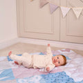 A baby in a light outfit smiles and raises an arm while lying on the Quilted Playmat, with cream cabinets and pastel bunting visible in the background.