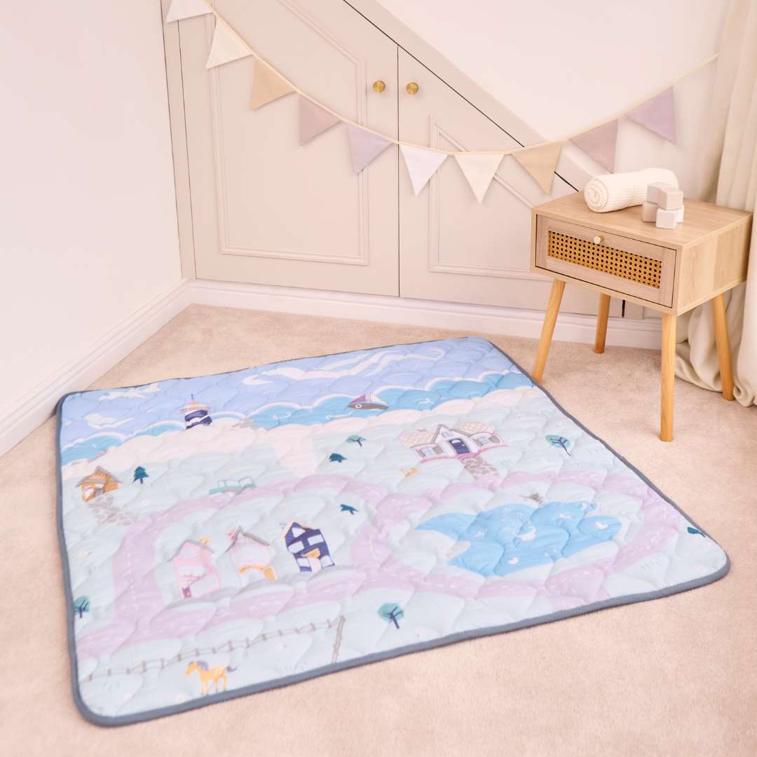 The Quilted Playmat, featuring pastel houses and clouds, is spread on a beige carpet near a wooden table and cream cabinet. Bunting above adds to the cozy, playful vibe of the space.