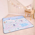 The Quilted Playmat, featuring pastel houses and clouds, is spread on a beige carpet near a wooden table and cream cabinet. Bunting above adds to the cozy, playful vibe of the space.