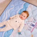 A light-skinned, red-haired baby in a cream bear-themed outfit lies on the Quilted Playmat, which features blue quilting with house, swan, and bear illustrations.