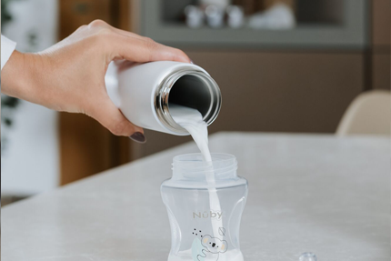 A hand pours milk from a stainless steel bottle into a Nuby UK baby bottle on a light countertop, demonstrating the Formula Making Bundle—an efficient feeding solution.