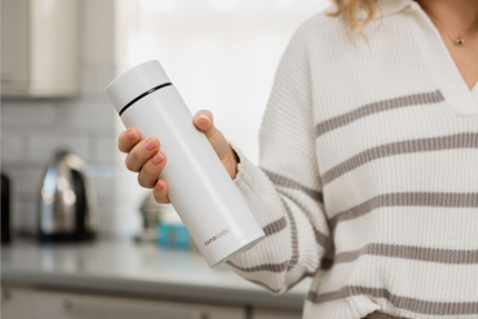 A person wearing a white and gray striped sweater holds a sleek Formula Making Set by Nuby UK in a modern kitchen, with the brand name printed near the bottom.