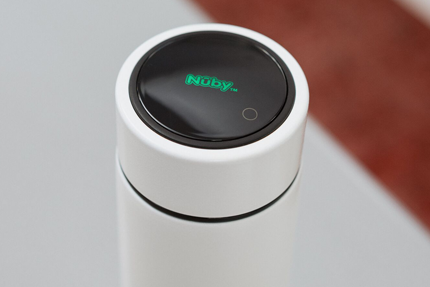 A close-up of a white cylindrical container with a black lid featuring the green Nuby UK logo and a small touch button, part of the Formula Making Bundle—a modern feeding system—on a light surface with a blurred brown background.