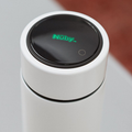 A close-up of a white cylindrical container with a black lid featuring the green Nuby UK logo and a small touch button, part of the Formula Making Bundle—a modern feeding system—on a light surface with a blurred brown background.
