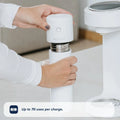 A person places the white, cylindrical Nuby UK RapidSterilise Baby Bottle Maker Steriliser onto its matching container. Text on screen: Up to 70 uses per charge. The product is displayed on a light countertop.