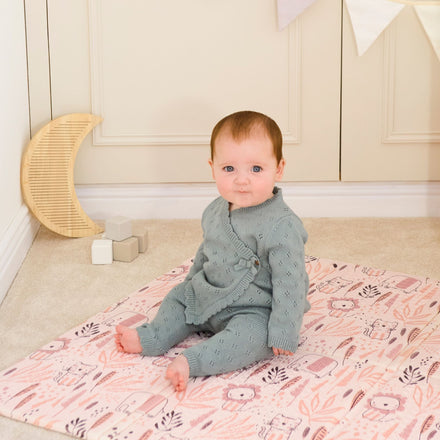 A baby in a light blue knitted outfit sits on the Fold N Go Play Mat with animal illustrations, surrounded by wooden blocks, a crescent moon decoration, and white bunting hanging on the wall.
