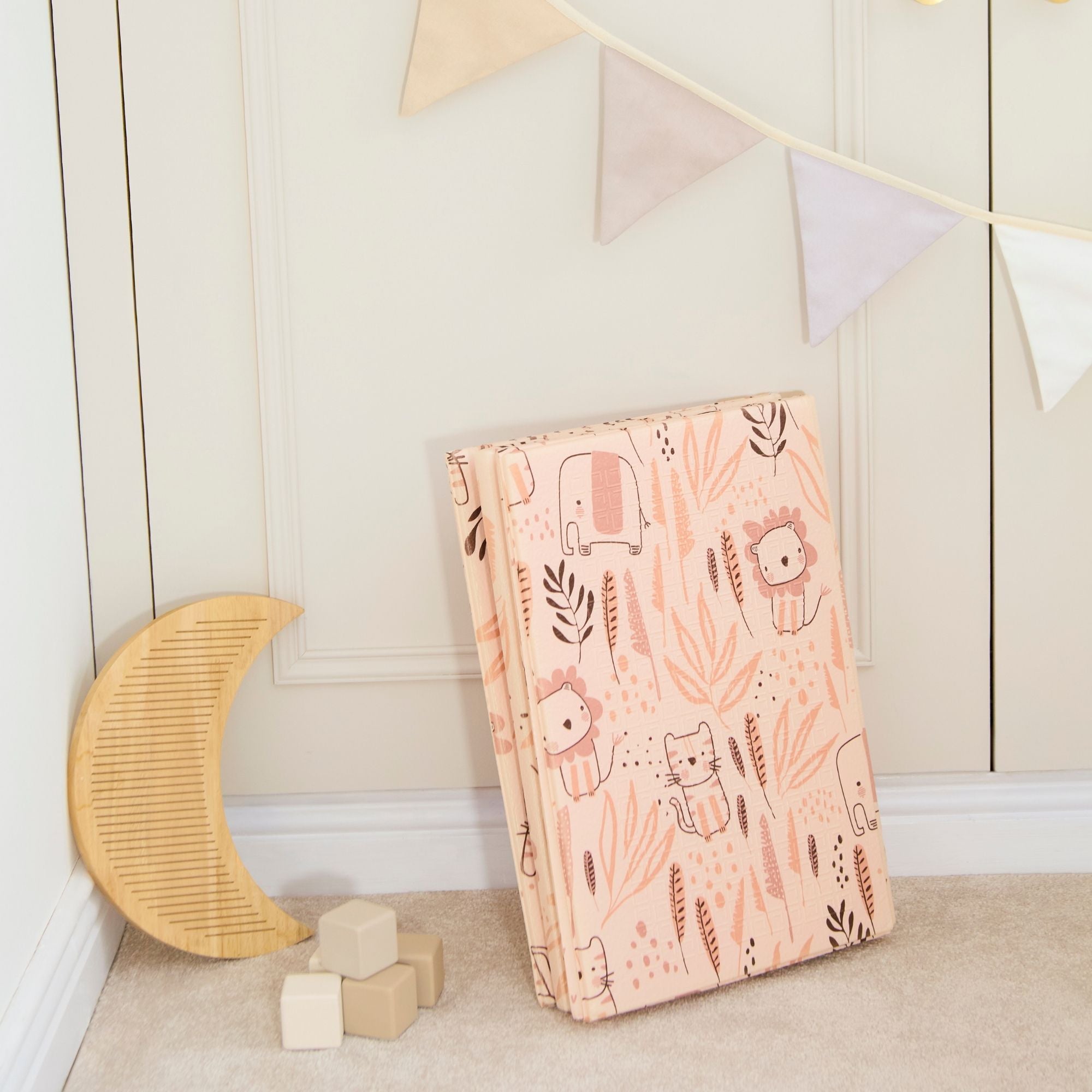 The Fold N Go Play Mat in pink with animal prints leans against the wall. Nearby, a wooden crescent moon and soft blocks rest on the carpet, while pennant banners decorate the wall above.