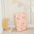 The Fold N Go Play Mat in pink with animal prints leans against the wall. Nearby, a wooden crescent moon and soft blocks rest on the carpet, while pennant banners decorate the wall above.