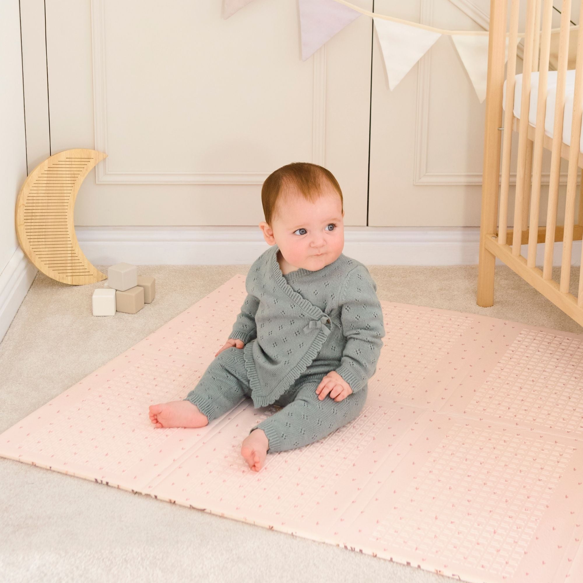 A baby in a green knit outfit sits on the Fold N Go Play Mat in a nursery with a crib, wooden blocks, a crescent moon decoration, and pastel bunting on the wall.