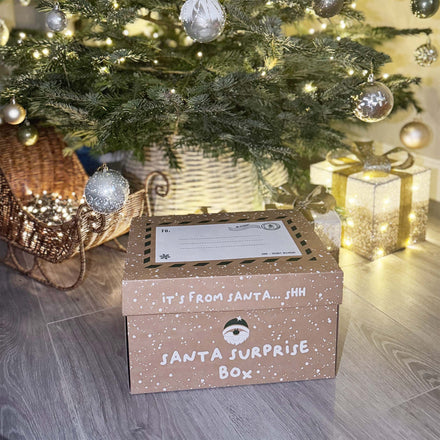 The Nuby Santa Surprise Box by Nuby, sat in front of a Christmas tree with gifting and light around it.