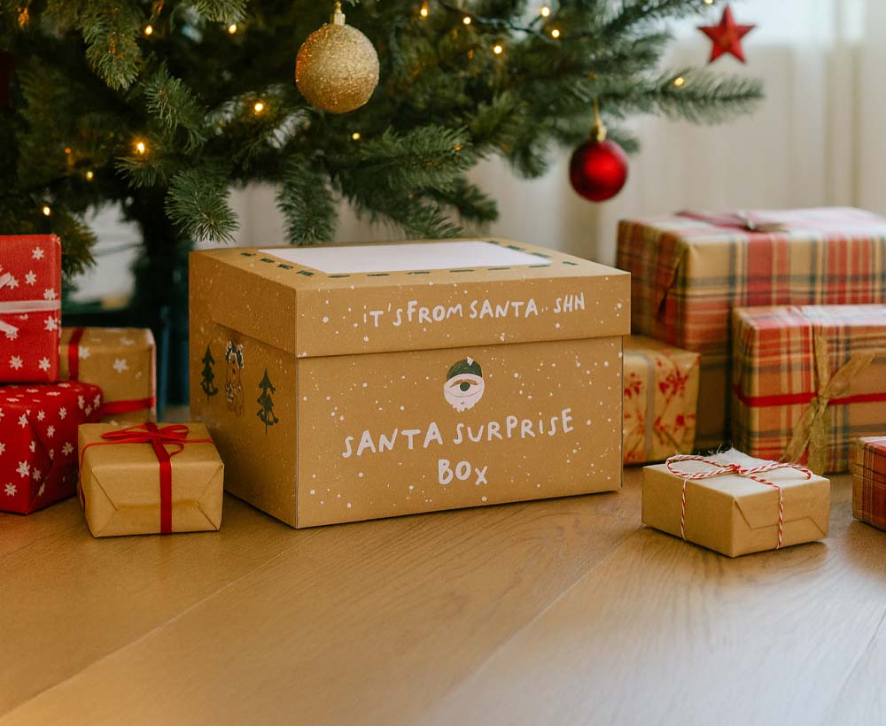 A festive Nuby UK Santa Surprise Christmas Bundle, labeled "Santa Surprise Box" with "It’s from Santa. Shh" on the lid, sits under a tree surrounded by baby gifts for ages newborn to 18 months.