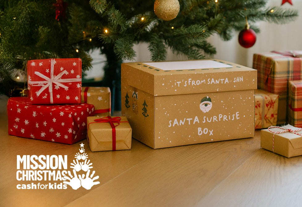 A photograph of the Santa Surprise Box from Nuby, laid underneath a Christmas tree surrounded by gift boxes and with the Mission Christmas Cash For Kids logo alongside it.