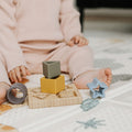 A young child in a light pink outfit sits on a patterned play mat, playing with the Nuby Shape Sorter Learner Toy—colorful silicone stacking toys and geometric blocks on a wooden board. The child’s face is not visible.